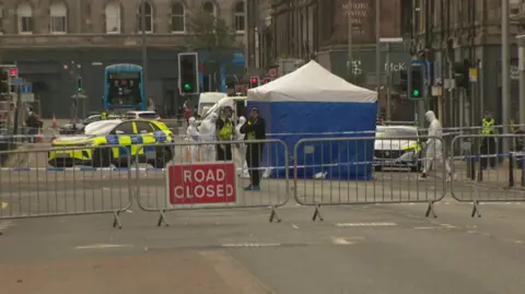 Forensic officers and police officers around a tent in the middle of a closed-off road