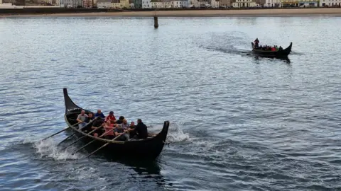 Two brown Viking longboats sail next to each other in the sea. Two teams of ten on board pull oars to row.