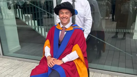 Family handout A man in a graduation gown sits in a wheelchair with his hands folded in his lap. He is looking at the camera and smiling.
