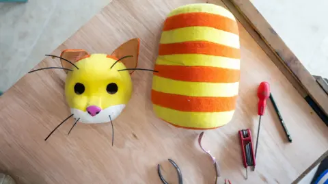 Eden Project The automaton cat is in pieces laid out on a table and pictured from above. The yellow head is separate to the orange and yellow striped body and there are various tools laid next to the parts, including a screwdriver and a pair of pliers.