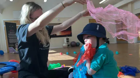 New school principle Katie Hankins sits cross legged on the floor with a little boy. Katie wears black leggings and a black t-shirt with 'Kiri Golding Theatre Arts' written on it in white. The little boy wears a light blue top with a collar and dark blue bucket hat that straps around the chin. They are playing with colourful scarves in pink, red and blue. In the hall behind them there is a trampoline and large rubber balls.