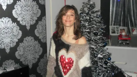 A woman with wavy shoulder length dark hair and an off the shoulder wooly jumper with a red love heart on the front stands smiling in front of a Christmas tree.