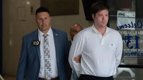William Duplessie wearing a white dress shirt is being led by police detective who is gripping his arm. 
