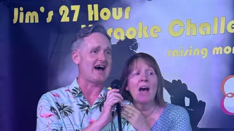 A man and a woman singing into the same microphone stand. "Jim's 27 hour karaoke challenge" is written in the background.