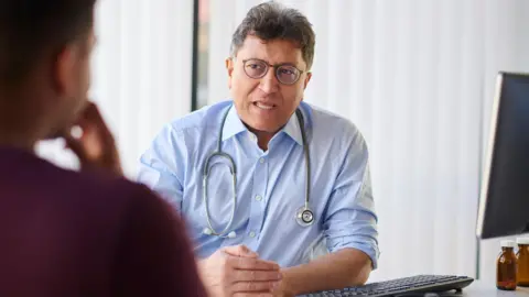 A doctor speaks to a seated patient across a desk in a bright consultation room, with a computer, keyboard, and small medicine bottles visible nearby.