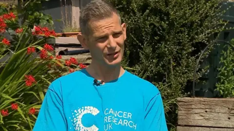 A grey-haired Adam Alderson wearing a blue Cancer Research UK t-shirt while being interviewed by the BBC in a sunny garden