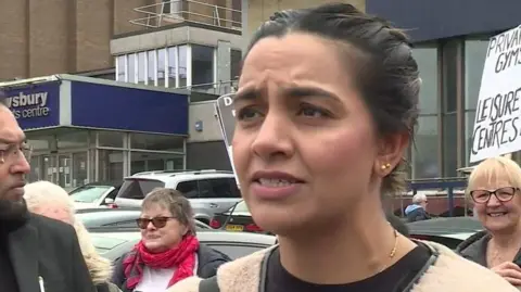 Olivia Richwald/BBC A woman with tied-up dark hair and gold earrings, stood in front of campaigners with placards.