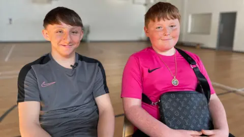 Two boys sitting in a sports hall - one wearing a grey t-shirt and the other is wearing a pink top, and a bag across his body