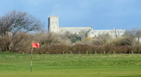 Des Blenkinsopp / Geograph A telephoto shot of Christchurch Priory in the distance, with a putting green and an orange flag of Solent Meads gold course in the foreground. The priory church is a huge stone medieval building with a square tower at one end.