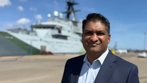 Andy Meeson/BBC Indy Wijenayaka is wearing a navy suit jacket over a pale blue shirt. The ship is behind him in the background.