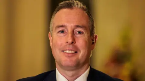 PA Media Paul Givan  is wearing a black suit jacket, white shirt and red tie.