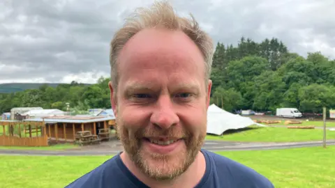 Ross Parkhill has short blonde hair and is wearing a navy shirt and is standing near a festival camping site.