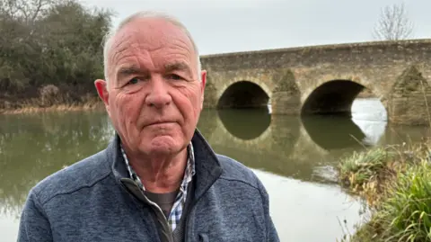 Ben Schofield/BBC Gerry Sansom standing outside looking down the camera. He has grey hair, blue eyes, and is wearing a casual blue, zipped top, over a jumped and a checked shirt. Behind him is a river and an old-looking stone bridge. Two of the bridge's arches can be seen, through which the greeny-grey water of the river is flowing. In the foreground on the right are some reeds, reaching down to the water's edge. Behind Gerry and to the left of frame is the far bank of the river, on which are trees and bushes. 