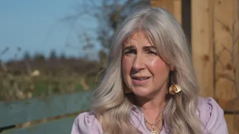 Nicola Welsh has long blond hair. She is wearing a lilac top and has large gold earrings and a gold chain with a heart symbol on it