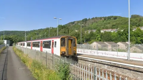 Google Abercynon train station. A white train with red doors is pulled up at the platform, with a metal fence on the other side of the tracks. A hillside covered in trees can be seen behind it.