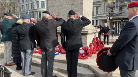 Rhys Thomas/BBC A general view of the Cenotaph in Jersey. Veterans are seen from behind. They are wearing suits and saluting. Wreaths are laid at the bottom of the cenotaph.