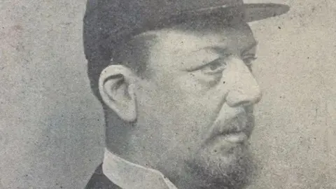 Leeds Museums A black and white portrait photograph showing a man in side profile. He has a beard and is wearing a smart, structured shirt and a flat-topped hat.