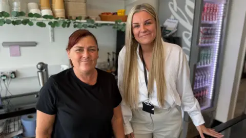 Donna Jones Employee Kerry Rowe is standing alongside owner Donna Jones, they are both behind the counter with a drinks dispenser in the back and they are both smiling into the camera.