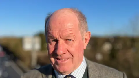 Tim Passmore smiles at the camera while standing on a bridge over the A14 carriageway. He is largely bald with some grey hair. He wears a grey suit jacket with a green jumper, shirt and tie underneath.