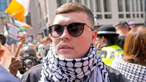 PA Media An up close image of Liam Óg Ó hAnnaidh, who is wearing a scarf and sunglasses. There is a crowd of people behind him.