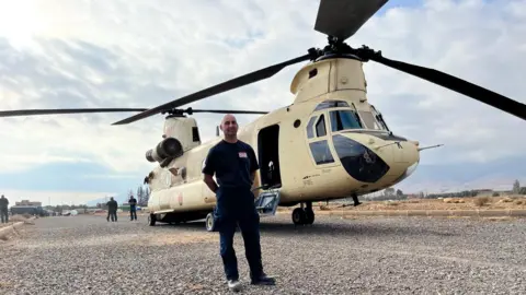 West Midlands Fire Service A man stands in front of a helicopter on a broad gravel path. The man man is bald and wearing a black t-shirt and trousers. The helicopter is cream with black blades.