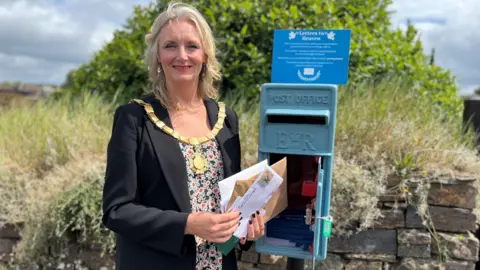A woman wearing a mayoral chain opens a bright blue postbox