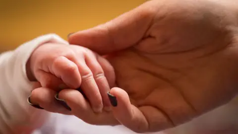 A small baby hand holding on to the fingers of an adult's hand. They have dark brown nails.