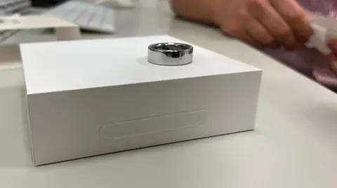 A metallic smart ring displayed on a white box on a desk, with a keyboard and a person’s arm blurred in the background.