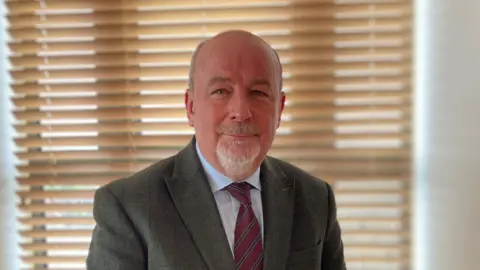 A man in a green tweed jacket is looking directly at the camera. He has very short hair at the sides and has a grey goatee beard. The man, who is also wearing a blue shirt and red tie, is sat in front of a window with horizontal wooden blinds, which are closed.