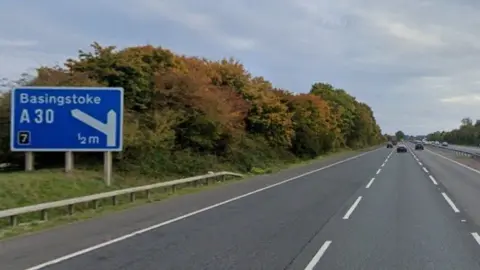 Three lanes of carriageway with trees and hedging to the left and blue motorway sign