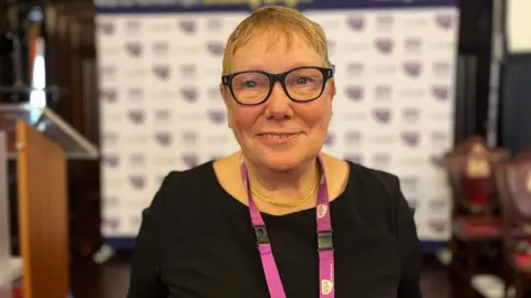 Sue Freeth. She has short hair and vision glasses with black frames is standing in the middle of the shot and looking into the camera. She is wearing black and has a lanyard on her neck with charity's logo. The promotional banner with charity's logo is in the background, that is blurred.