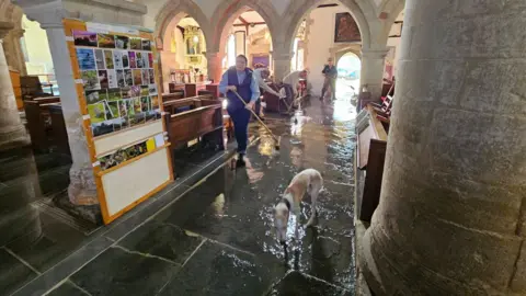 Leila Mather Four people with brooms sweeping water out of a medieval church while a dog paddles around in the shallow water that covers the stone floor.