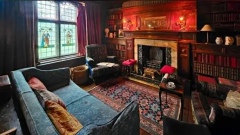 Ian Hughes A richly decorated room with a large fireplace, sofas, stained glass on some of the window panes and a decorative rug on the floor