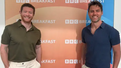 Matthew Carter and Lawrence Price stand infront of a BBC Breakfast board as they smile at the camera. Both have short hair and beards.