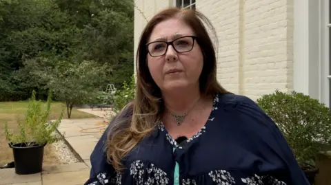 Ruth Horton looks slightly off-camera. She has long dark hair and is wearing a blue dress or blouse and glasses. She is outside in a garden with plants and the exterior wall of a house visible behind her.