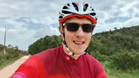 Victor Thompson A man wearing sunglasses, a bicycle helmet and red lycra smiles into the camera. He is outdoors and is cycling on a dusty road with green bushes and overgrowth on each side of the road.