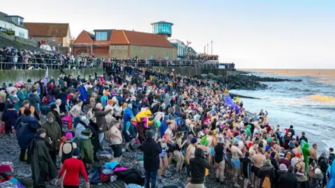 Hundreds of people on a beach. Some people are wearing their swimsuits while other people are wearing big winter coats. They are all facing the north sea. There are even more people onlooking by the pier. 