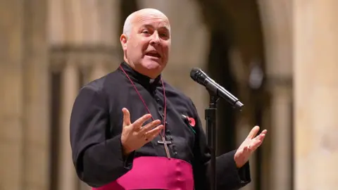 Getty Images Archbishop of York, Steven Cottrell, speaks to a gathered congregation in York Minster during a vigil calling for peace in Israel and Gaza on 11 November 2023. He is a bald man with white hair on the side of his head, his hands are outstretched. He is wearing a black cassock with a pink waist sash, and there is a metal cross on a chain around his neck.