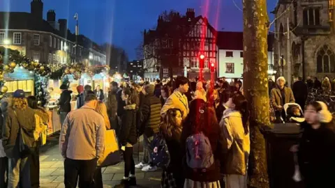 Tens of people stand around York Christmas Market at night time.