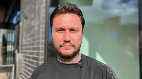 Joe standing outside his block of flats. He has a beard and short dark hair. He is wearing a black top.  