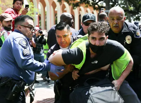 Getty Images Police officers hold a man wearing a mask and hi viz jacket at a protest on USC