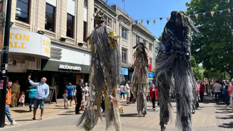 Emma Baugh/BBC Three stilt walkers move through a town centre. They wear grey outfits that are covered in long threads of fabric.