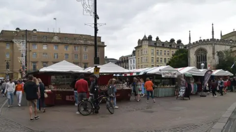 NChadwick/Geograph The photo is of the Cambridge market. The sky is grey and cloudy and there are buildings of various colours in the background. On the ground the camera is focused on the market stalls which have white, blue, yellow or green striped tarp covers atop. The stalls are on cobbled flooring and around the market is a smoother concrete path. There are customers walking around the market, one with a bike, browsing the stalls. Signs for a olive stall and a coffee stall can be seen clearly from the camera angle.