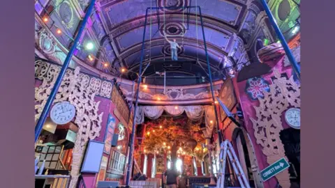 Richie Hurren A man stands at height on trapeze equipment in a colourful Victorian venue with soft pink, purple and red lighting.