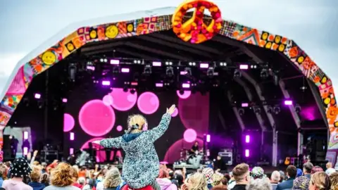 Crowds gather in front of the Camp Bestival main stage which is lit with pink spotlights. The arched canopy over the stage has a CND logo at the top. A small child is sitting on someone's shoulders and waving their hands in the air.