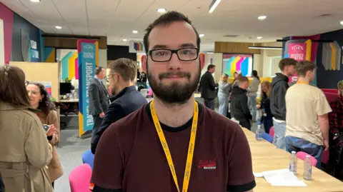 Student lawyer Liam Lennon-Swales is looking straight at the camera. he is wearing a burgundy T-shirt, spectacles and a yellow lanyard. He has a black beard and black hair.