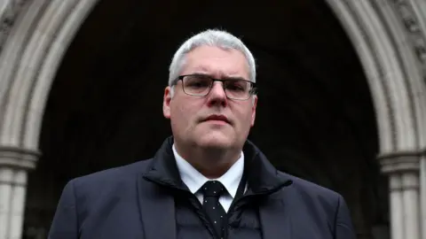 PA Media Gavin Robinson is looking down the barrel of the camera. Hew has grey hair and black glasses. He is wearing a white shirt, black tie and black coat.