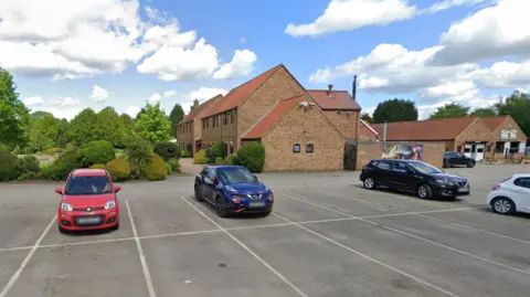 Google Parking in a rural hotel. In the photo you can see a group of cars parked, between empty spaces and with the backdrop of some hotel buildings, all of them with sloping roofs and some trees.