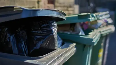 A close-up image of a black rubbish bin with bin-bags in. There are two green bins behind which are also full.