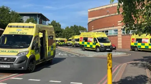 Six ambulances outside a hospital building. The vehicles are all mainly yellow with green rectangle patterns on. The hospital building is red brick and the lower part has a curved shape. 
